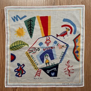 Puede incluir: Pañuelo cuadrado, blanquecino, con un diseño abstracto y colorido. Presenta una forma geométrica central con la palabra "SILENCE" y una figura. Incluye el texto "MAISON DE FOUS" y "Henry Miller Museum of Art".