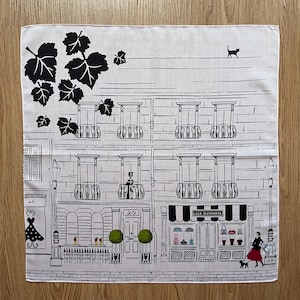 May include: A white cotton bandana featuring a black and white illustration of a row of buildings with a shop called "Lulu Guinness" on the right. The illustration includes a black cat on the roof, a woman in a red dress, and a small dog.