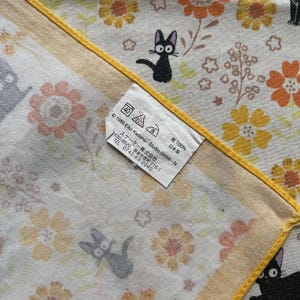 May include: A white and yellow floral patterned handkerchief with black cat illustrations. The handkerchief has a yellow border and a tag with washing instructions and Japanese text.