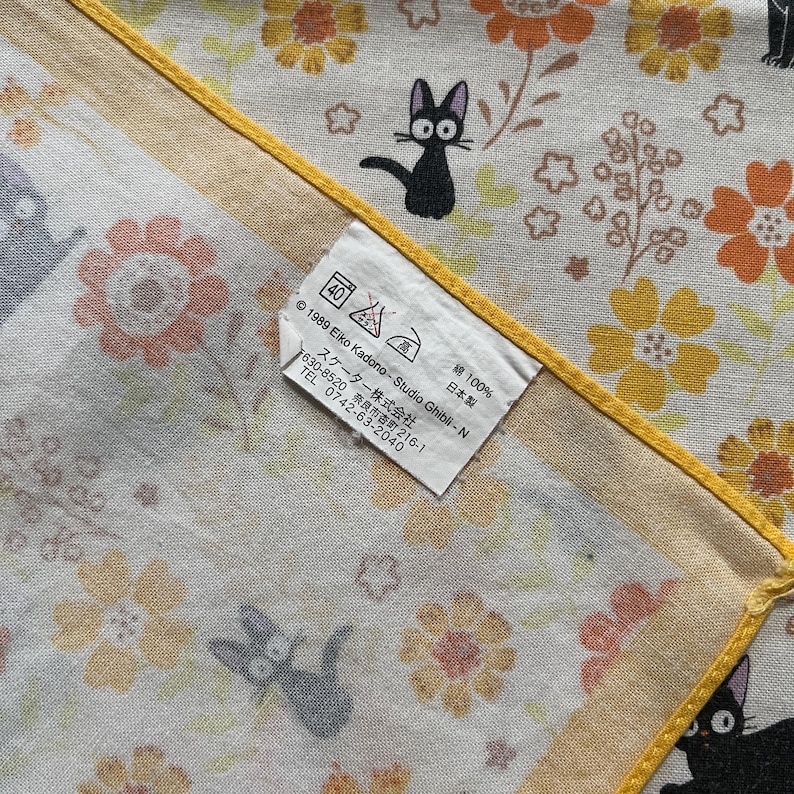 May include: A white and yellow floral patterned handkerchief with black cat illustrations. The handkerchief has a yellow border and a tag with washing instructions and Japanese text.