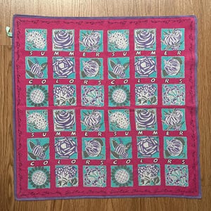 Puede incluir: Un pañuelo cuadrado con fondo rosa vibrante y una cuadrícula de cuadrados turquesa con ilustraciones florales moradas y blancas. El texto "SUMMER COLORS" está impreso en blanco en el centro y "United Colors" bordea el borde.