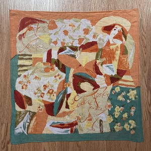 May include: Square scarf with an abstract illustration in shades of orange, yellow, and teal. The design features a woman in a hat, floral patterns, and the handwritten text "Jackie". The scarf has a teal border.