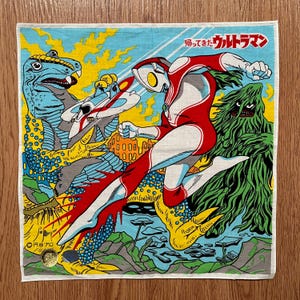 May include: Square, vintage-style handkerchief featuring a colorful illustration of a superhero in a red and white suit, battling monsters. The design includes Japanese text and a retro aesthetic, with a blue, yellow, and green color scheme.