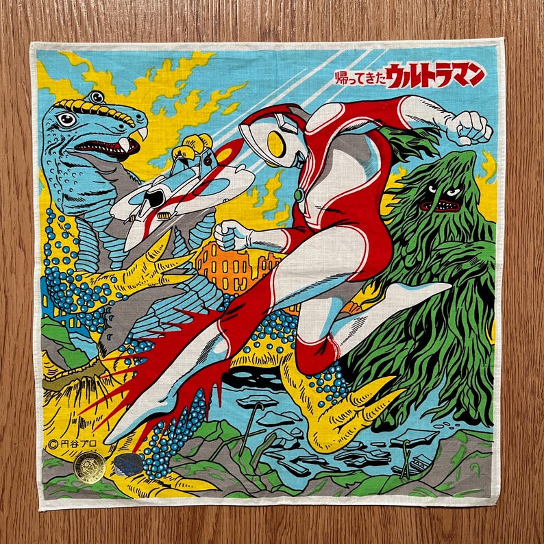 May include: Square, vintage-style handkerchief featuring a colorful illustration of a superhero in a red and white suit, battling monsters. The design includes Japanese text and a retro aesthetic, with a blue, yellow, and green color scheme.