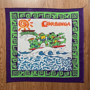 Könnte beinhalten: Ein lilafarbenes Bandana mit grünem Rand, auf dem die Teenage Mutant Ninja Turtles auf einem Surfbrett reiten, mit den Worten "Cowabunga" und "Teenage Mutant Ninja Turtles" bedruckt.