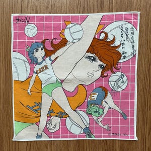 May include: Pink and white square handkerchief with a volleyball anime design. The handkerchief features illustrations of volleyball players, volleyballs, and Japanese text. The background has a grid pattern.