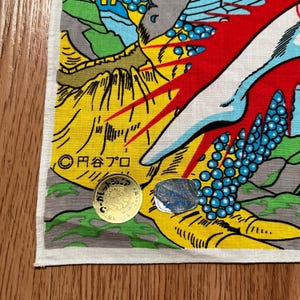 May include: A colorful Japanese-style textile with a dragon design. The fabric features yellow, red, blue, and green hues, with Japanese text and a gold coin. The design includes a dragon's claw and a mountain landscape.