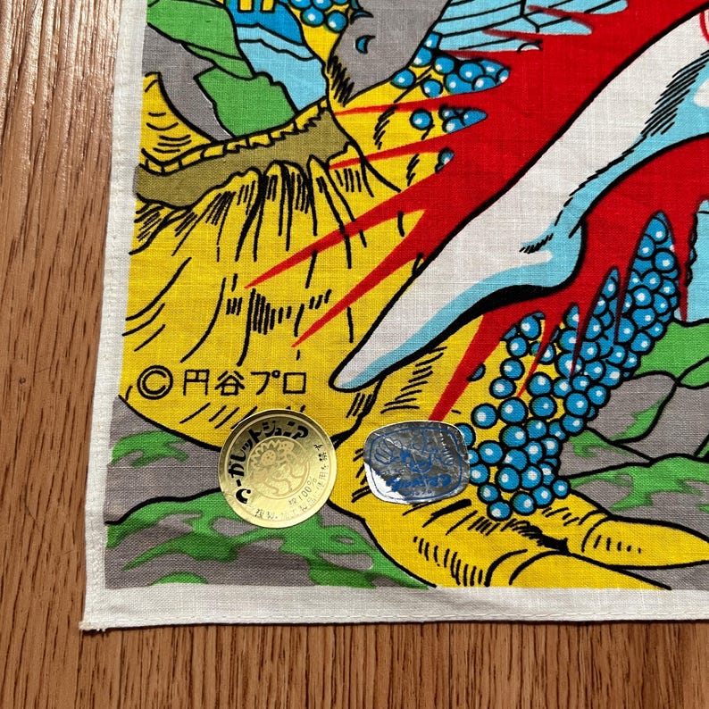 May include: A colorful Japanese-style textile with a dragon design. The fabric features yellow, red, blue, and green hues, with Japanese text and a gold coin. The design includes a dragon's claw and a mountain landscape.