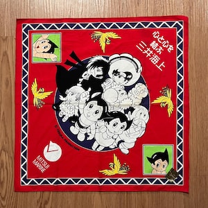 May include: Red bandana with a white and black illustration of Astro Boy and his friends. The bandana has a blue border with a white diamond pattern. The text "Mitsui Marine" and "Tezuka Production" are printed on the bandana.
