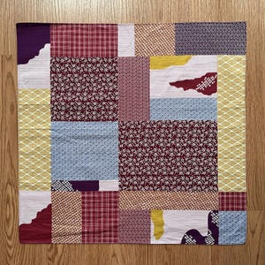 Op de afbeelding: Een patchwork textiel met een verscheidenheid aan patronen en kleuren. Het ontwerp bevat vierkanten van rode ruit, witte bloemen en geometrische vormen in tinten paars, geel en blauw. Het textiel is waarschijnlijk een decoratief item.