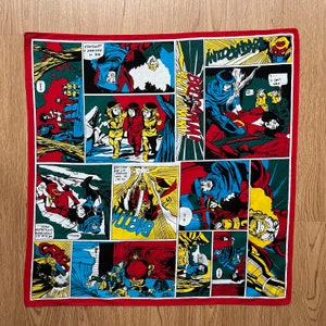 May include: A red bandana with a comic book style print featuring characters in various scenes. The bandana has a white border and includes the text "TRAATAROOUMY" and "ANCIENT F OF POWER".