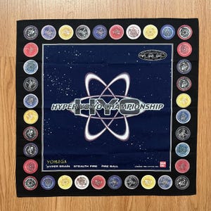 May include: Black square bandana with a navy blue centre featuring the text "HYPER YO-YO CHAMPIONSHIP" and a white graphic. The border is black with colourful circular designs. The bandana also has the text "YOMEGA" and "HYPER BRAIN STEALTH FIRE FIRE BALL".
