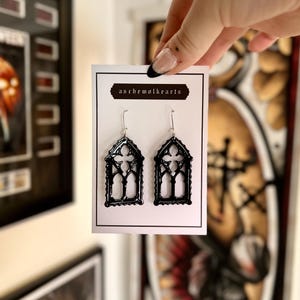 Puede incluir: Pendientes negros con forma de ventana gótica con ganchos plateados. Los pendientes se muestran en una tarjeta blanca con el texto "aschemalkrarts". Los pendientes son sostenidos por una mano.