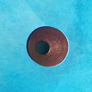 May include: A copper-colored, circular object with a smaller, dark circle in the center, set against a bright blue background. The copper circle has a textured, speckled appearance. The contrast between the warm copper and cool blue creates a striking visual.