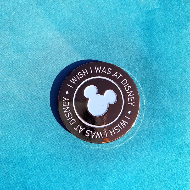 May include: A round, metallic pin with the text "I Wish I Was At Disney" encircling a light blue Mickey Mouse silhouette. The pin has a shiny, rose gold finish and is set against a turquoise background.