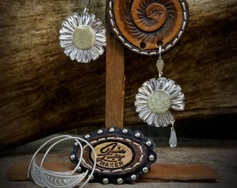 May include: Silver earrings with a daisy design and a dangling crystal. The earrings are displayed on a leather stand with the text "Janie's artistry maker".