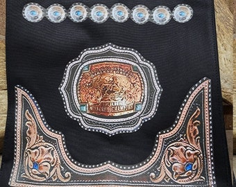 May include: Black canvas tote bag with a brown leather patch that says "Ginny Maker". The bag has a design of a silver belt buckle with rhinestones and a blue stone in the center. The buckle has the text "Bringing Home The Best" and "Darlene Green 19".