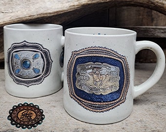 May include: Two white ceramic mugs with a brown leather buckle design. The mugs feature a blue and gold design with the text "100 Miles One Day Western States Trail Ride".