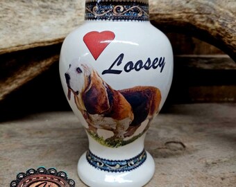 May include: White ceramic urn with a red heart and the name "Loosey" written in black. A basset hound dog is pictured on the urn. The urn has a blue and gold trim around the top and bottom.