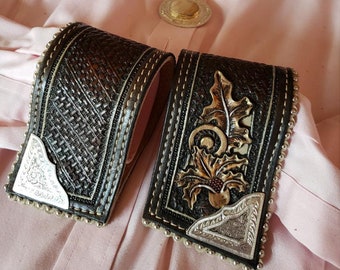 May include: Two brown leather cufflinks with silver accents. One cufflink features a woven pattern and the other has a floral design. The cufflinks are on a pink fabric background.