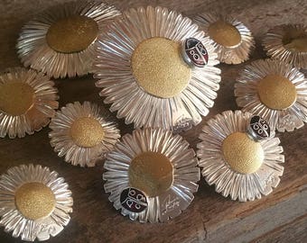 May include: A collection of silver daisy-shaped brooches with gold centers and black ladybug accents. The brooches are arranged on a wooden surface.