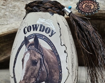 May include: A white ceramic vase with a brown horse illustration and the word "Cowboy" on the front. The vase is decorated with a braided horsehair tassel.