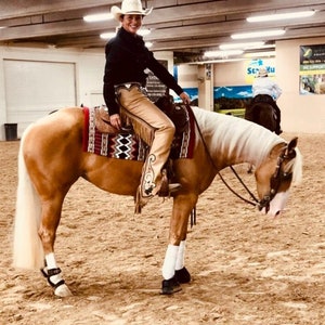 Puede incluir: Una mujer con un sombrero de vaquero blanco y polainas de cuero marr&oacute;n monta un caballo palomino en un recinto interior. El caballo tiene una crin y una cola blancas.