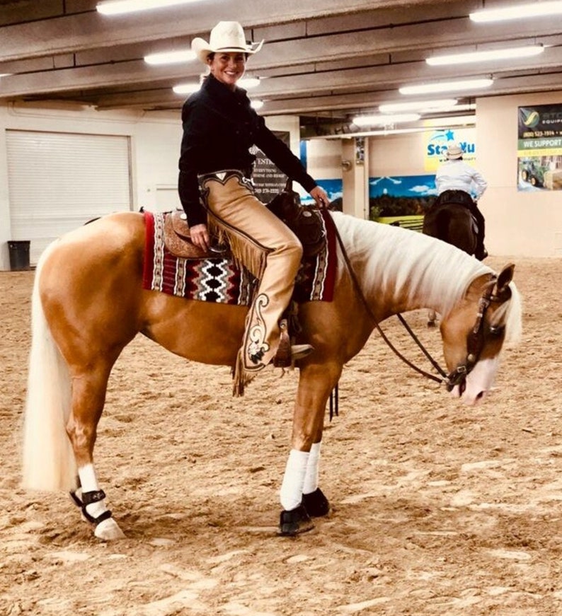 Puede incluir: Una mujer con un sombrero de vaquero blanco y polainas de cuero marr&oacute;n monta un caballo palomino en un recinto interior. El caballo tiene una crin y una cola blancas.