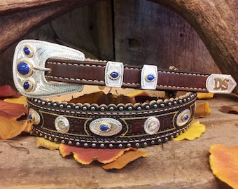 May include: A brown leather belt with a silver buckle and blue gemstone accents. The belt has a decorative design with silver and gold accents. The belt buckle has the initials "DS" on it.