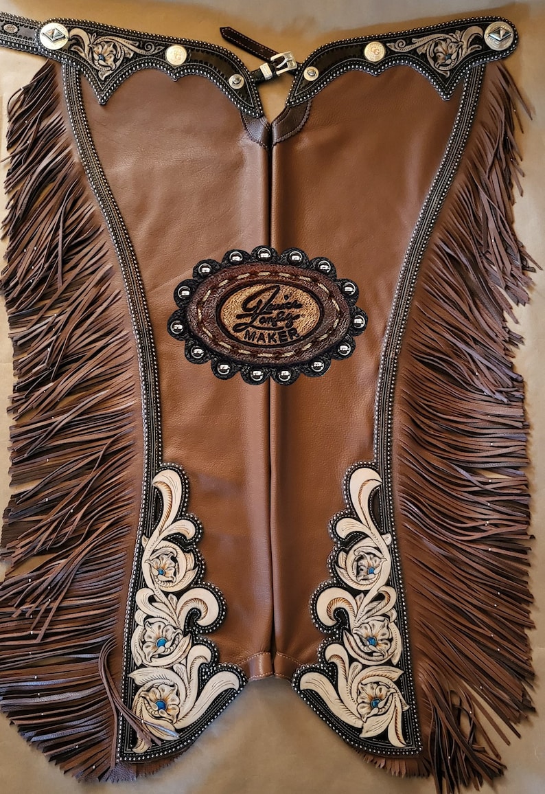 Puede incluir: Chaps de cuero marr&oacute;n con flecos, bordados florales blancos y una insignia ovalada plateada con el texto "Janice Long Maker".