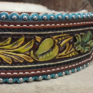 Puede incluir: Un collar de cuero marr&oacute;n con detalles en turquesa y plata. El collar est&aacute; decorado con un dise&ntilde;o floral y tiene un peque&ntilde;o charm redondo de plata y turquesa con el texto "Jamie Lady Maker".