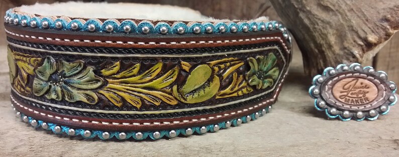 Puede incluir: Un collar de cuero marr&oacute;n con detalles en turquesa y plata. El collar est&aacute; decorado con un dise&ntilde;o floral y tiene un peque&ntilde;o charm redondo de plata y turquesa con el texto "Jamie Lady Maker".