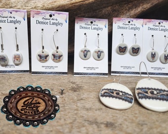 May include: A set of handmade ceramic earrings featuring various designs, including butterflies, cows, and dogs. The earrings are displayed on cards with the artist's name and website information. The cards are on a wooden surface with a leather tag that says "Denice Langley Maker".