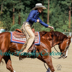 Puede incluir: Una mujer con una camisa azul y pantalones marrones monta un caballo marr&oacute;n. El caballo lleva una silla de montar con una manta de silla de montar con estampado azul, rojo y blanco. La mujer sostiene las riendas en su mano derecha. El caballo est&aacute; en movimiento y la mujer se inclina hacia adelante.