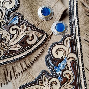 Puede incluir: Primer plano de un par de pantalones de cuero con intrincados dise&ntilde;os tallados que representan p&aacute;jaros y motivos florales. Los pantalones est&aacute;n decorados con conchos plateados con piedras de vidrio azul.