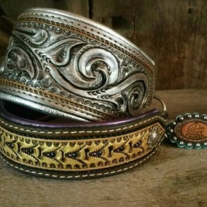 Puede incluir: Dos collares de perro de cuero con intrincados dise&ntilde;os en relieve. El collar superior es plateado con un patr&oacute;n floral y el collar inferior es marr&oacute;n con un patr&oacute;n floral dorado y costuras moradas. El collar inferior tiene una hebilla plateada y un amuleto turquesa con el texto "Linda's Leather Maker".
