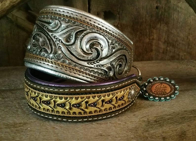 Puede incluir: Dos collares de perro de cuero con intrincados dise&ntilde;os en relieve. El collar superior es plateado con un patr&oacute;n floral y el collar inferior es marr&oacute;n con un patr&oacute;n floral dorado y costuras moradas. El collar inferior tiene una hebilla plateada y un amuleto turquesa con el texto "Linda's Leather Maker".