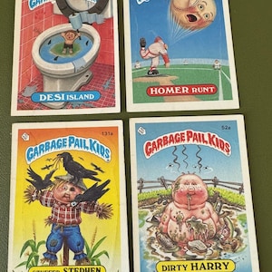 May include: Four Garbage Pail Kids trading cards featuring different characters. The cards are in a light blue, yellow, and white color scheme. The characters are a boy in a toilet, a baseball player, a scarecrow, and a pig in mud. The cards have the text "Garbage Pail Kids" on them.