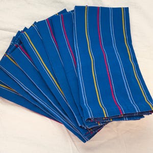 May include: A stack of blue cloth napkins with vertical stripes in white, yellow, red, and light blue. The napkins are fanned out, showcasing the striped pattern. The background is a plain, off-white fabric.