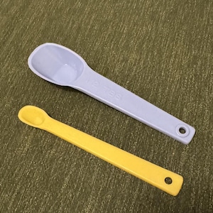 Peut inclure: Deux cuillères à mesurer en plastique. L'une est violet clair et marquée "1/2 TBS". L'autre est jaune. Les deux cuillères ont un trou dans le manche pour être suspendues. Les cuillères sont sur une surface verte texturée.