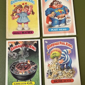 May include: Four Garbage Pail Kids trading cards featuring different characters. The cards are white with a colorful border and a picture of a character on the front. The characters are Double Heather, Nat Nerd, Grilled Gil, and Richie Retch. The cards are from the 1987 Topps Garbage Pail Kids series.