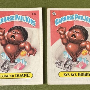 May include: Two Garbage Pail Kids trading cards. The card on the left features a cartoon baby with brown skin, wearing a nappy, and stuck in a toilet. The card is titled "Clogged Duane". The card on the right features a cartoon baby with brown skin, wearing a nappy, and stuck in a toilet. The card is titled "Bye Bye Bobby".