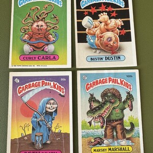 Op de afbeelding: Vier vintage Garbage Pail Kids ruilkaarten. De kaarten bevatten stripfiguren met humoristische en groteske thema's. De personages zijn onder meer Curly Carla, Bustin' Dustin, Beth Death en Marshy Marshall. De kaarten hebben een witte rand en kleurrijke achtergronden.