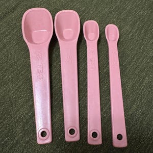 Peut inclure: Ensemble de quatre cuillères à mesurer roses. Les cuillères indiquent les mesures suivantes : 1/2 cuillère à soupe, 1 cuillère à café, 1/4 cuillère à café et 1/8 cuillère à café. Chaque cuillère a un trou dans le manche.