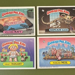 May include: Four vintage Garbage Pail Kids trading cards. The cards feature cartoon illustrations with humorous themes. One card reads "Welcome Matt", another "Dinah Saur", a third "Target Margaret", and the last "Nailed Neil".