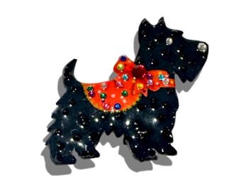 May include: A black Scottish Terrier brooch with a red and orange bow and collar. The brooch is embellished with colorful rhinestones and small black accents. The dog is standing on a white surface.