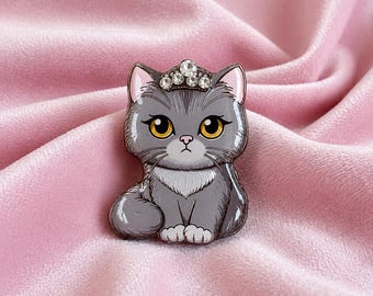 May include: A gray cat-shaped enamel pin with a sparkling rhinestone tiara. The cat has yellow eyes and is sitting with its paws together. The pin is set against a pink, velvety background.