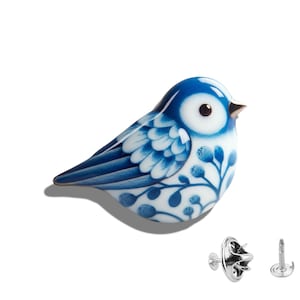 May include: A decorative ceramic bird figurine in blue and white, featuring a floral pattern. The bird has a shiny surface and is shown with two silver-coloured pin backs, possibly for use as a brooch or lapel pin.