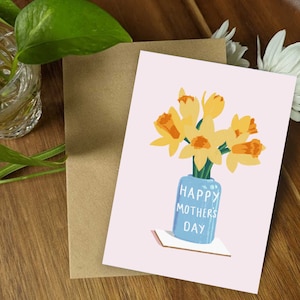 May include: A pink card with a bouquet of yellow daffodils in a blue vase. The vase has the text "Happy Mother's Day" on it.