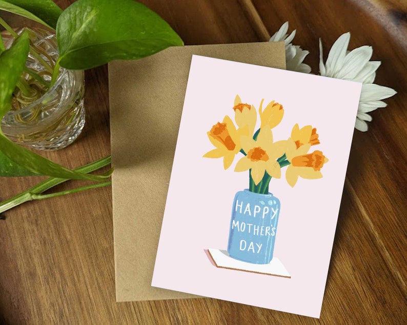 May include: A pink card with a bouquet of yellow daffodils in a blue vase. The vase has the text "Happy Mother's Day" on it.
